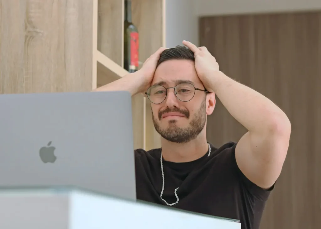 A man frowns and places his hands on his head while looking at a laptop. 