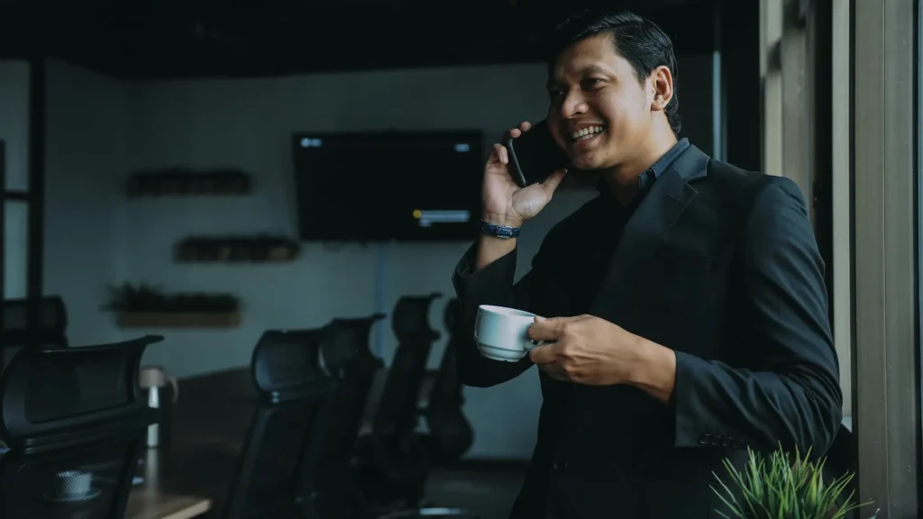 A man speaking on the phone in a meeting room while holding a small white cup.